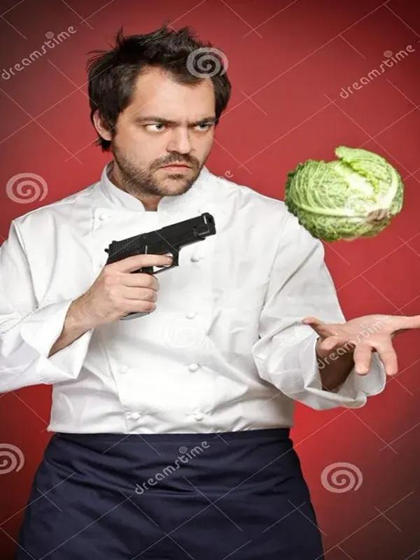 A humorous stock photo of a man dressed as a professional chef against a solid red background. He wears a white chef's coat and a dark blue apron, with a tense, angry expression on his face. In his right hand, he holds a black handgun, pointing it directly at a whole head of green Savoy cabbage that appears to be floating or tossed in the air just above his open left palm. The entire image is overlaid with faint Dreamstime watermarks.